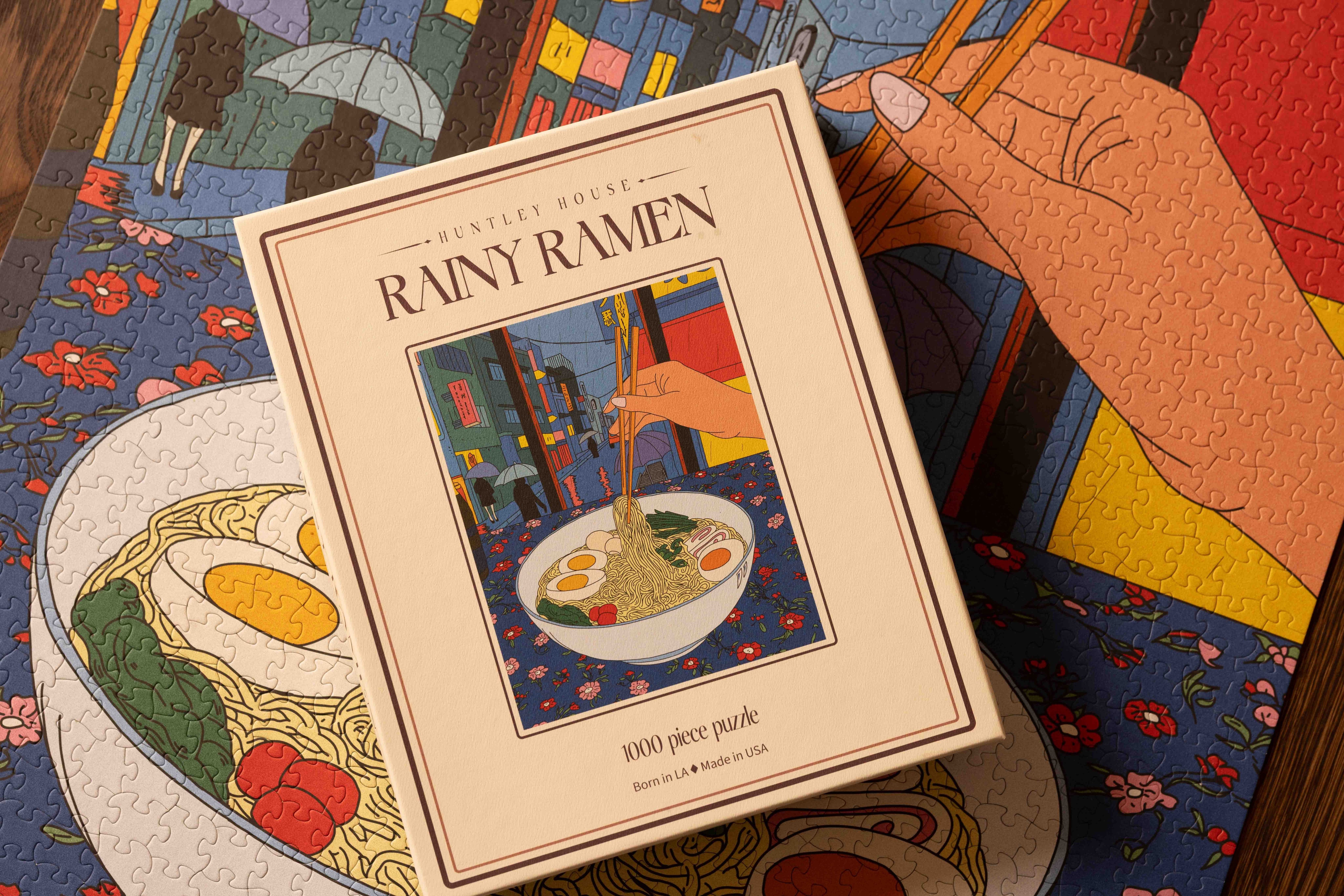 A lifestyle view of the Huntley House Rainy Ramen puzzle box placed on top of the colorful ramen-themed jigsaw puzzle pieces