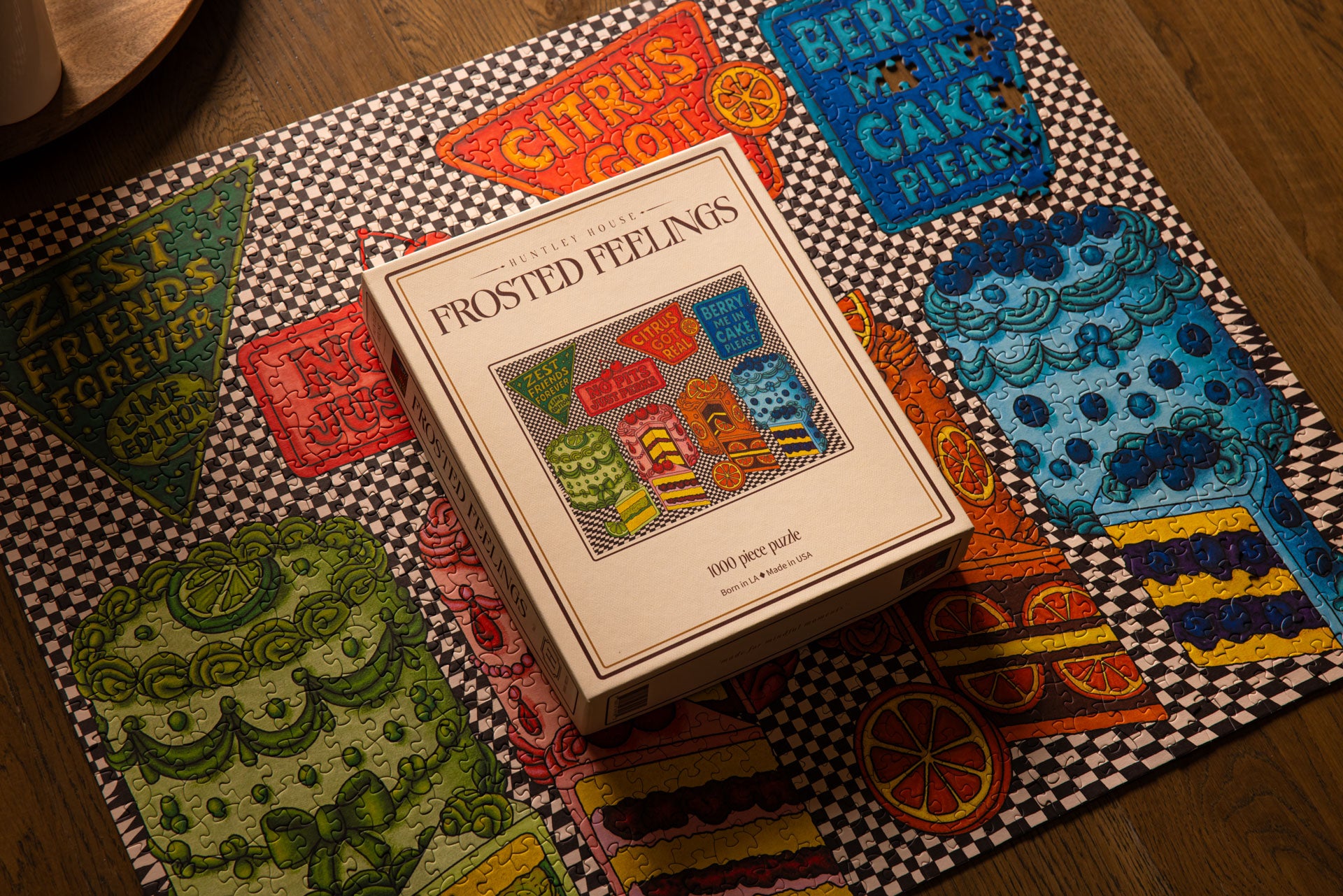 The Huntley House Frosted Feelings 1000 piece puzzle box placed on top of the fully assembled colorful cake and checkered pattern puzzle
