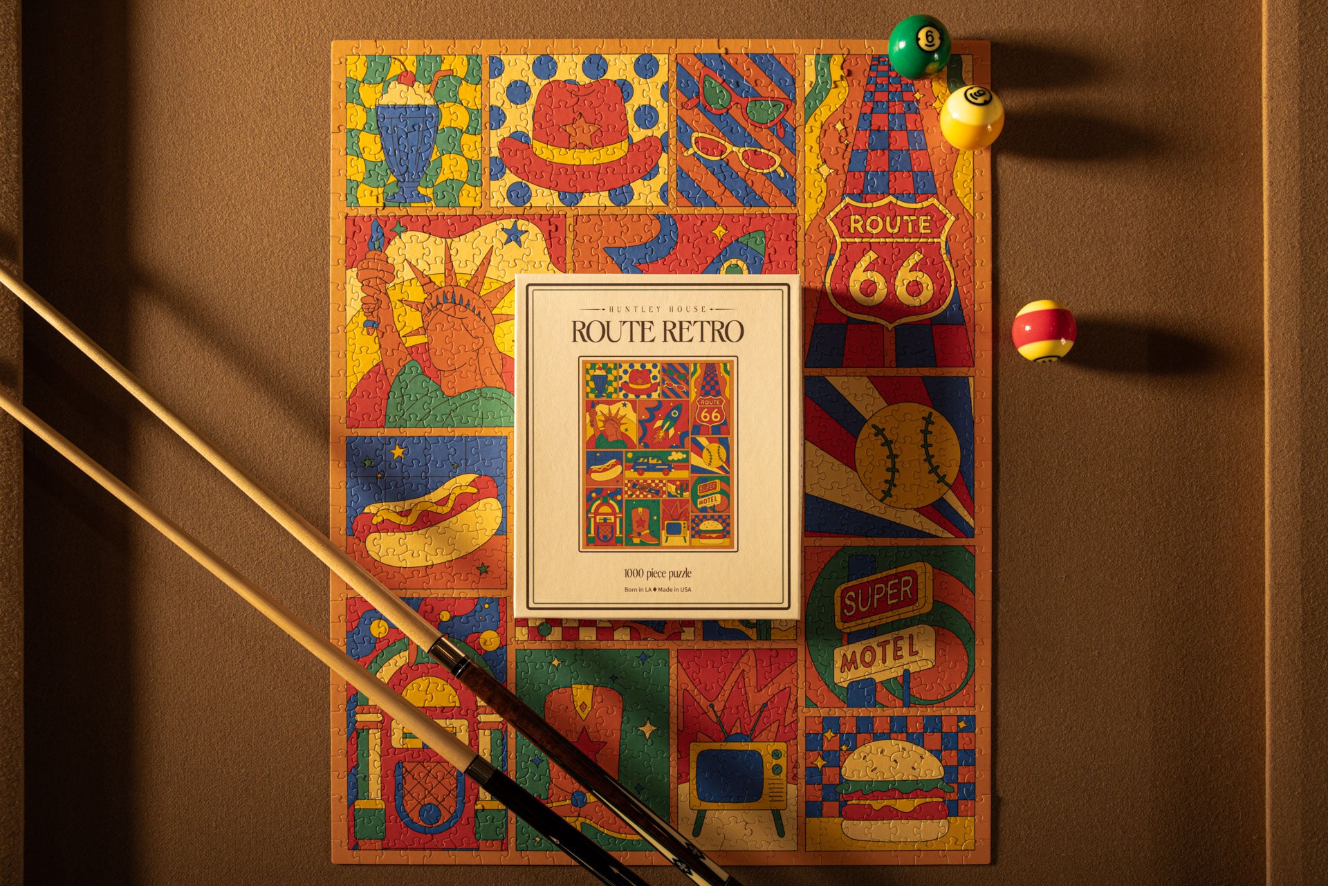 Fully assembled Route 66 Retro 1000 piece puzzle with its box and pool cues on a brown billiard table surface